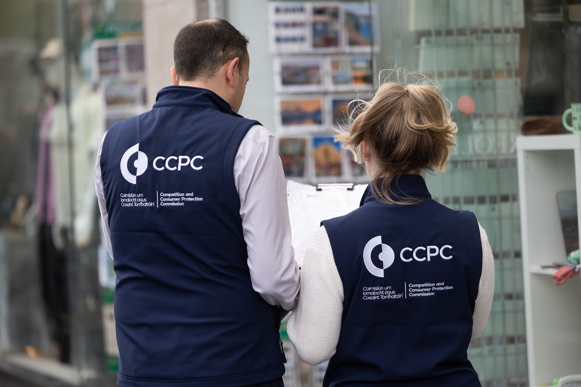 Two officers in CCPC-branded gilets with their backs to the camera, examining a clipboard outside a shop