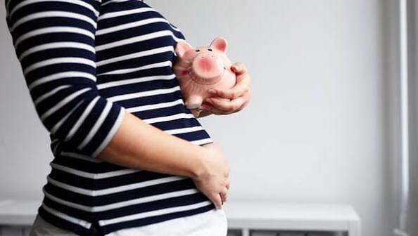 A pregnant person cradles their stomach and a pink piggy bank