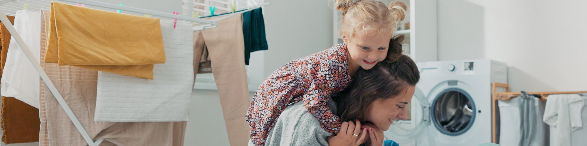 A child climbs on a woman's back while they are in a laundry room with a tumble dryer in the background
