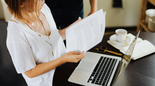 A woman reviews a contract printed on paper, with her laptop open
