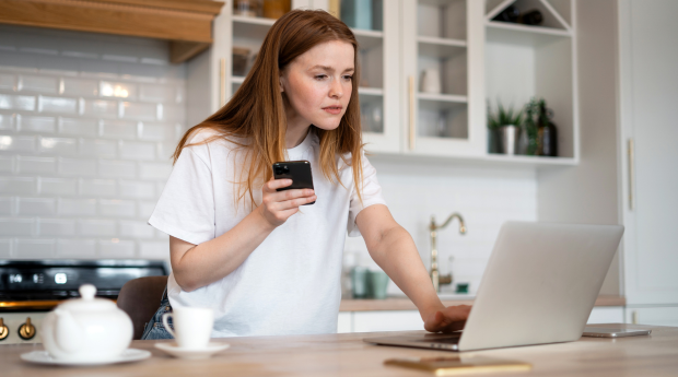 A woman in a kitchen looks at her laptop with a concerned expression with her phone in her hand