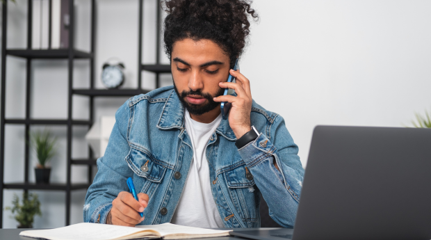 A young man is on the phone taking notes in his notebook with his laptop open
