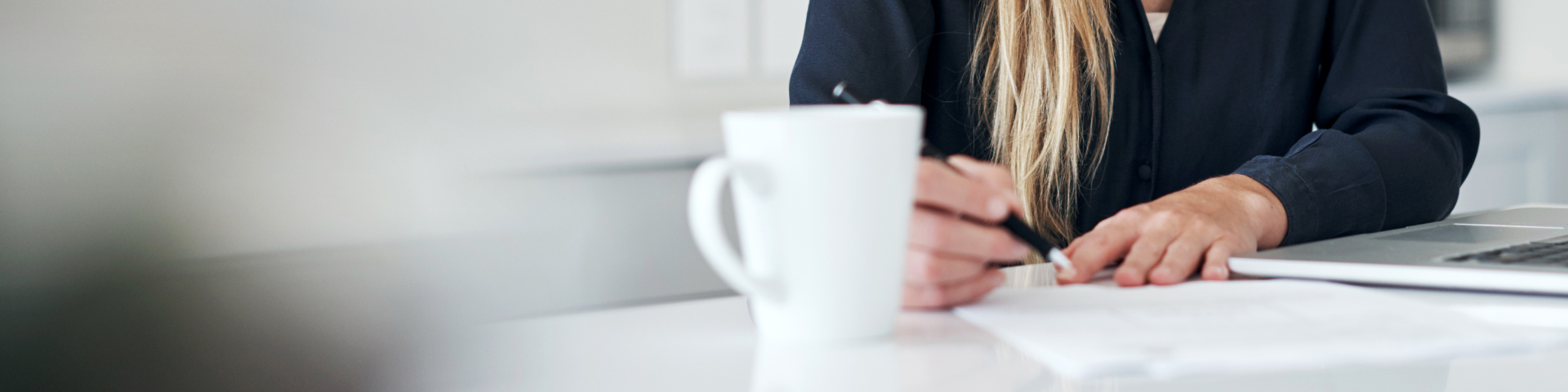 A woman fills out paper work with her laptop open and a mug on her desk