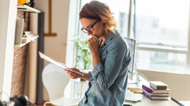 A woman in casual clothes leans against a table reading a piece of paper work and appears to be thinking deeply