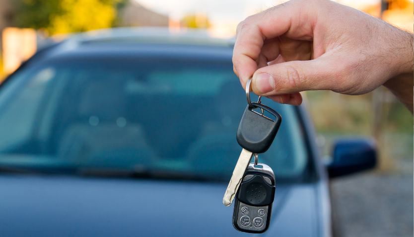 A hand holds a set of car keys with a car parked in the background