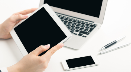 Hands holding a computer tablet while there is a laptop and mobile on a desk.