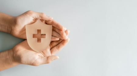 Two hands holding a wooden medical plus symbol within a shield.