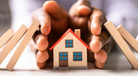 Hands surrounding a toy house while blocks fall either side of it.