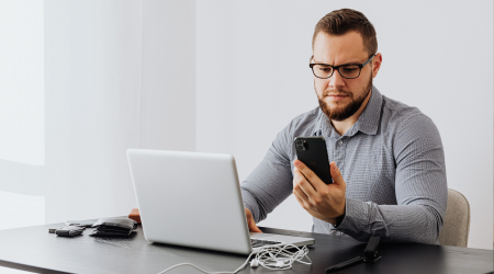A man sits at a desk looking at his phone. He has a laptop on the desk in front of him.