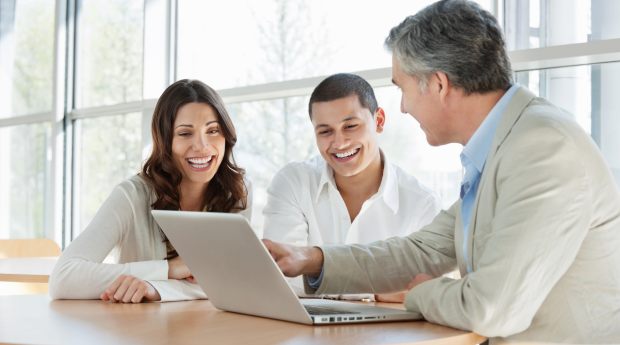 Three people smiling looking at a computer while the third person points to it.
