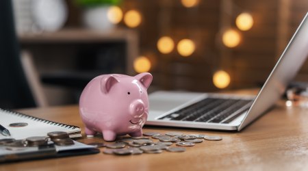 A pink piggy bank and a laptop sit in the middle of a desk. Scattered on the desk are coins.