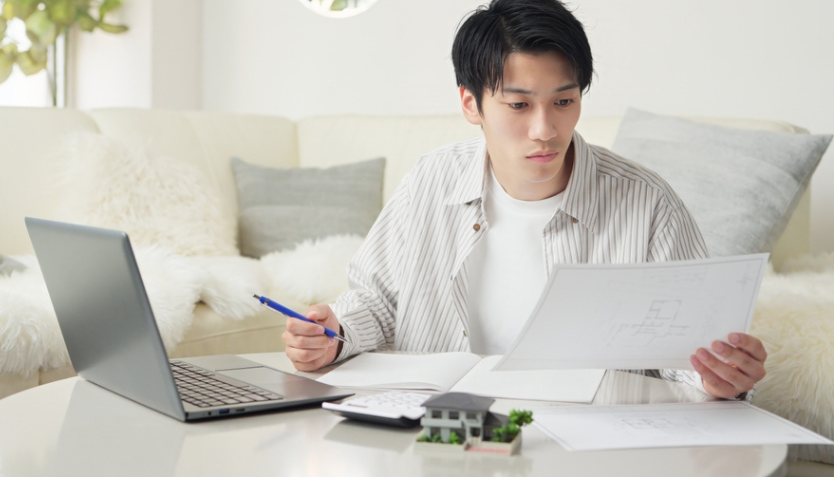 Man looking at a piece of paper and holding a pen, in front of an open laptop.